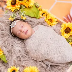 A peaceful newborn baby, swaddled snugly in a beige wrap, sleeping in a wooden crate adorned with bright yellow sunflowers.