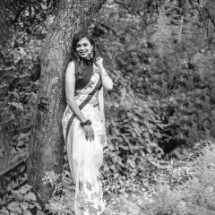 A classic black and white portrait of a woman in a saree, her smile radiating warmth against the rustic texture of a tree trunk.