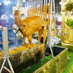 A full view of the whole roasted mutton on the spit, part of our grand barbecue setup with hanging seekh kebabs in the background.