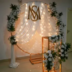 This modern and elegant backdrop was designed for a wedding anniversary. It features a circular frame with white drapes, fairy lights, and a custom monogram, creating a romantic and personalized setting.