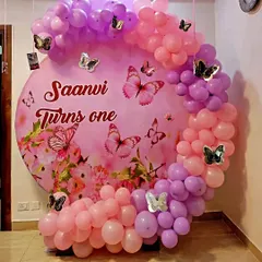 A lovely butterfly-themed first birthday decoration for "Saanvi". The design features a custom pink backdrop with the baby's name, surrounded by a beautiful arch of pink and purple balloons with decorative butterflies.