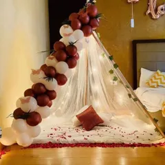We can bring the romance indoors with a cozy canopy decoration right in your bedroom. This setup features a sheer teepee with fairy lights, a rich maroon and white balloon garland, and scattered rose petals for an intimate celebration.