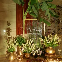 A traditional welcome setup featuring a clear acrylic sign, lush banana leaves, and elegant brass vases filled with tuberoses.
