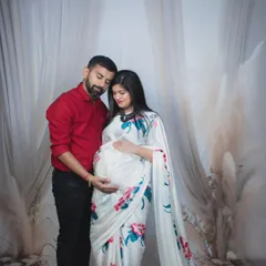 A tender moment between expecting parents. The father-to-be gently holds the baby bump as they stand together in a boho-themed studio setup.