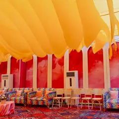 A bold and creative Haldi lounge area featuring colorful printed sofas set against a pink and red backdrop, all under a ceiling of flowing orange fabric waves.
