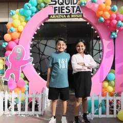 The happy birthday kids, Araina and Rish, posing in front of their 'Squid Fiesta' party entrance. I designed this colorful and fun entrance arch with balloons and themed cutouts to set an exciting tone for their Squid Game inspired celebration.