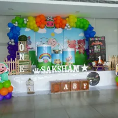 A vibrant Cocomelon setup for Saksham's first birthday. The decor features a rainbow balloon arch and a full cast of character cutouts, creating a playful and colorful scene.
