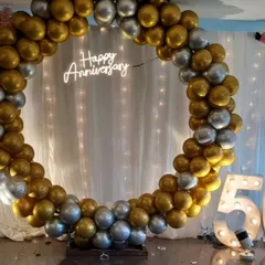 A modern and stylish 50th-anniversary decoration. This setup features a large circular balloon arch in gold and silver, a "Happy Anniversary" neon sign, and light-up marquee numbers.