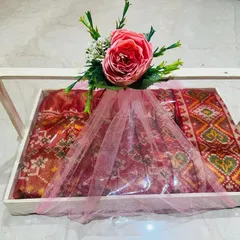 This is how I deliver a complete gift experience. A traditional Patola-style saree is carefully packed in a wooden tray with a clear acrylic top, wrapped in soft pink tulle, and finished with a floral arrangement, ready for any shagan ceremony.