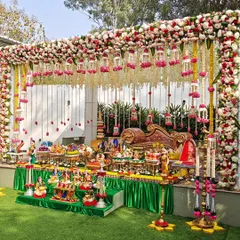 An outdoor baby shower setup featuring a stunning floral canopy with hanging arrangements. The multi-tiered display includes traditional dolls and an abundance of offerings, creating a festive and natural setting.