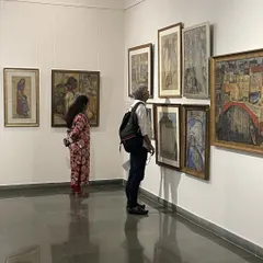 Two visitors examining a dense salon-style hanging of Vidya Bhushan's paintings. This curatorial choice allows for a comparative study of his works from different periods.
