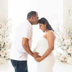 An intimate and tender moment between a couple. The partner gently kisses his wife's forehead in a bright, all-white setting filled with beautiful white floral arrangements.