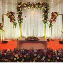 This modern stage design combines a lush floral arch with industrial-style hanging bulbs against a clean white curtain. The setup is framed by matching floral stands, offering a chic and sophisticated look for an engagement or reception.
