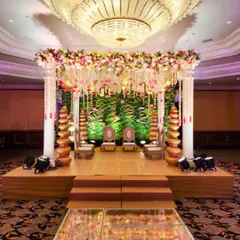 This indoor mandap features a unique glass aisle filled with flower petals leading to the stage. The backdrop is a cascade of floral strings against a lush green wall, creating a garden-like feel indoors.