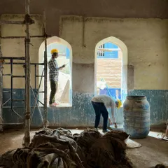 Workers carefully restore the interior of a classroom. The image shows the layers of old paint and plaster being stripped back to reveal the original wall surface, the first step in understanding the building's history.