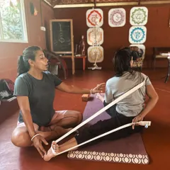 We teach you how to use props to make yoga accessible and effective for every body. Here, an instructor guides a student in Janu Sirsasana (Head-to-Knee Pose) using a strap to create gentle traction and protect the lower back.