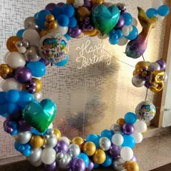 A mermaid-themed balloon ring in a mix of blue, purple, gold, and silver. The design features a mermaid tail, heart-shaped foil balloons, and a "Happy Birthday" neon sign.