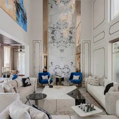 This view highlights the symmetry and scale of a double-height living room in our Chennai project. A book-matched marble wall serves as a dramatic backdrop, flanked by bold cobalt blue armchairs that create an intimate seating nook within the larger space.