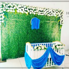 A simple and elegant naming ceremony setup with a modern white crib. The backdrop is a lush green wall topped with white flowers, with blue drapes adding a pop of color.