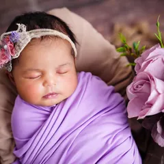 A close-up of a sleeping newborn wrapped in a lovely lavender swaddle, adorned with a floral headband and surrounded by matching flowers.