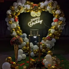 A glamorous birthday backdrop featuring a large heart-shaped balloon arch in gold and white. A "Happy Birthday" neon sign glows in the center, with a pedestal for the cake, all set for a memorable celebration.