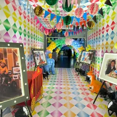 An immersive carnival-themed hallway decorated with diamond-patterned walls and flooring. The entrance is lined with lit-up photo frames on easels, creating a personal gallery for the guest of honor.