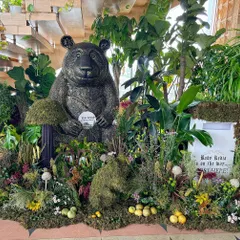 A whimsical woodland setup for a baby shower, featuring a large, friendly bear sculpture surrounded by moss, ferns, and a mailbox for "post" to the baby.