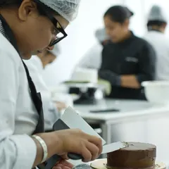 A student getting a smooth, sharp finish on a chocolate ganache cake. You will learn how to properly crumb coat and apply a final layer of frosting, a fundamental skill for professional-looking cakes.