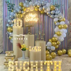 Here is a sophisticated ring balloon decoration I did for a 10th birthday. The setup includes a full arch of metallic gold, silver, and white balloons, a holographic curtain backdrop, an illuminated number '10', and marquee letters spelling the name 'SUCHITH'.