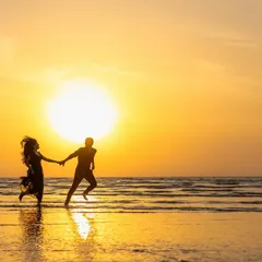 A classic sunset silhouette of a couple running hand-in-hand along the water's edge. The golden light and their joyful movement create a feeling of freedom and romance.