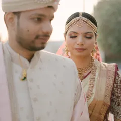 A soft, romantic portrait of a couple on their wedding day. The groom's classic cream sherwani with a light pink stole creates a gentle and elegant look beside his bride.