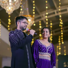 The groom gives a heartfelt speech during the Sangeet night, with his bride looking on. The backdrop of warm string lights creates an intimate setting.