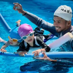 Learning with friends makes it fun. Here, young swimmers use pool noodles to practice their kicking technique together, supported and guided by their coach.