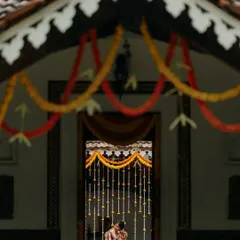 An artistic shot of the bride and groom, framed by the traditional architecture and floral decorations of a South Indian wedding venue. The composition creates a sense of intimacy and timelessness.