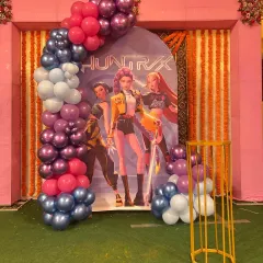 A 'Huntrix' themed party setup with a balloon arch in shades of purple, pink, and blue, set against a backdrop of marigold garlands.
