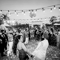 A wide-angle black and white shot of the couple dancing, surrounded by their friends and family under a canopy of string lights at a Goa wedding party.