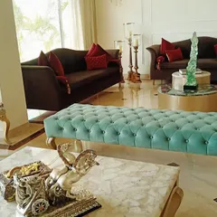 A wider perspective of a luxurious living room, centered around a custom-made tufted turquoise bench. The space also includes classic brown sofas with red accent cushions and a round coffee table, creating a balanced and opulent seating area.