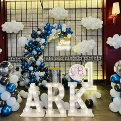 For a classy and simple first birthday, we designed this cloud-themed decor. The setup includes a balloon arch in shades of blue, black, and silver, marquee letters spelling the name 'ARK', and soft cloud-like balloon clusters to create a dreamy atmosphere.