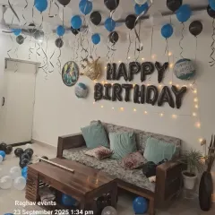 A classic birthday room decoration with blue, black, and white balloons on the ceiling. The setup includes 'Happy Birthday' foil letters, a crown foil balloon, and fairy lights.