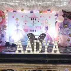 For a magical princess party, I designed this beautiful stage featuring a castle backdrop and two Disney princesses. The large, illuminated marquee letters for the name 'AADYA' create a perfect centerpiece for photos, complemented by soft pink and purple balloon decorations.