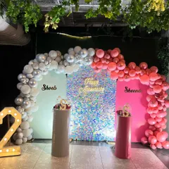 A personalized anniversary setup with a sparkling silver sequin wall. The decor includes two backdrops with the couple's names and balloon arches in coordinating silver and pink.