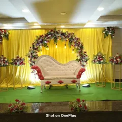 A reception stage with a bright yellow backdrop and a multi-colored floral arch. The classic red and white diwan adds a touch of traditional elegance to the vibrant setting.