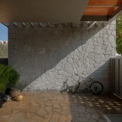 The entrance yard features a Sira stone wall and floor laid in a random rubble pattern. The diagonal shadow cutting across the textured surfaces sets the tone for the earthy, tactile experience of the home.