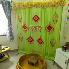 We can adapt our Haldi decor for any space, like this cozy living room setup. We used a light green curtain backdrop with traditional hangings, a wooden 'हळद' sign, and our signature golden Urli to create a festive corner.