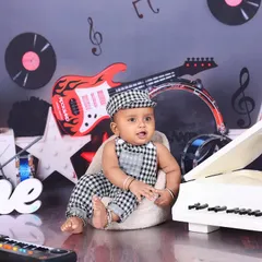 A baby boy sits at a miniature white piano in a rock-and-roll themed session. The guitars, drums, and musical notes on the wall create a fun and energetic atmosphere.