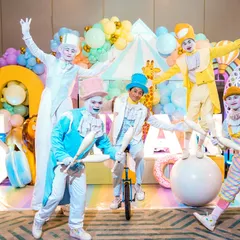A troupe of pastel-colored circus performers, including jugglers and a unicyclist, posing with the birthday boy at a grand carnival party.