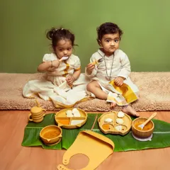 A heartwarming lifestyle shot for a kids' brand, featuring two toddlers using bamboo feeding sets. The traditional outfits and banana leaf placemat add a unique cultural touch, making the scene feel authentic and adorable.