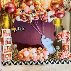 A charming first birthday setup with a baby elephant theme, using rose gold and pink balloons, "BABY" blocks, and a balloon-filled number '1'.