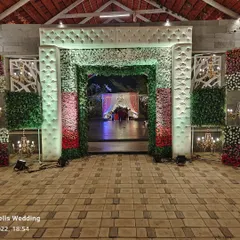 A grand main entrance designed for a wedding reception. The archway is covered in a mix of white, pink, and red flowers, framed by lush greenery and elegant white cushioned panels.
