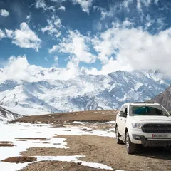 A white SUV is parked against a magnificent backdrop of snow-covered Himalayan peaks under a brilliant blue sky. This image perfectly captures the scale and beauty of a Spiti road trip.